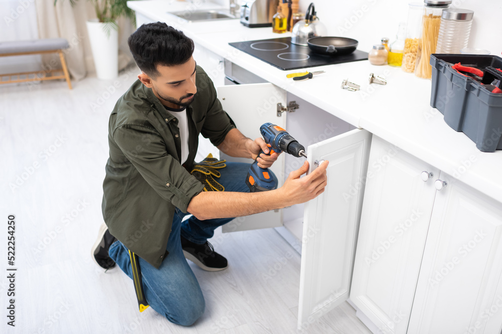 Wall mural Arabian man with electric screwdriver fixing cabinet under worktop in kitchen