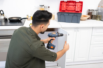 Young arabian man with electric screwdriver fixing door of kitchen cabinet near toolbox on worktop