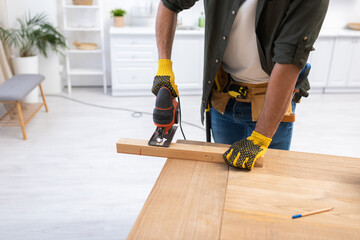 Cropped view of man in gloves holding jigsaw machine while sawing plank at home