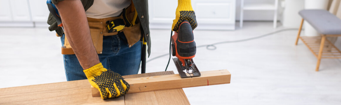 Cropped view of carpenter in tool belt sawing wooden plank at home, banner