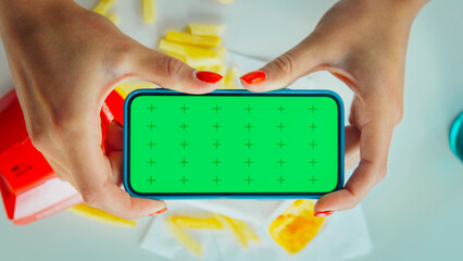 Female hands hold horizontal smartphone with chroma key green screen over table at cafe. French...