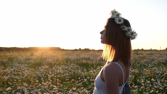 A beautiful girl is enjoying life in a daisy field at sunset. A young woman with a wreath of daisies on her head smiles happily in nature.
