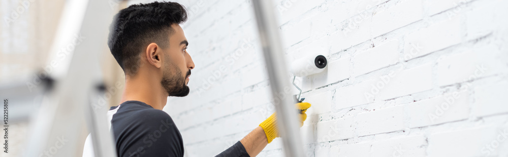 Wall mural side view of muslim man in glove coloring wall with roller near blurred ladder at home, banner