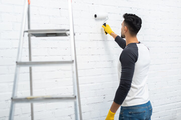 Arabian man in gloves coloring wall near ladder at home