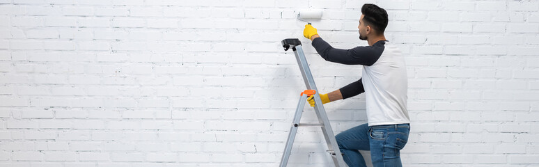 Muslim man in gloves coloring brick wall while standing on ladder at home, banner