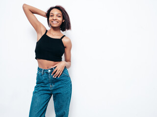 Fototapeta premium Portrait of young beautiful black woman. Smiling model dressed in summer jeans clothes. Sexy carefree female posing near white wall in studio. Tanned and cheerful