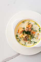 Norwegian red salmon fish soup with vegetables, herbs and cream, top view of a bowl of soup on a white marble background