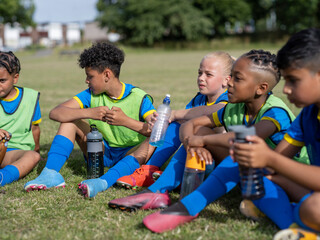 Children (8-9) dressed in uniforms sitting on soccer field - obrazy, fototapety, plakaty
