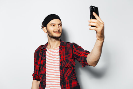 Studio portrait of young handsome smiling man making selfie photo with smartphone on white background. Wearing red casual clothes and black head band.