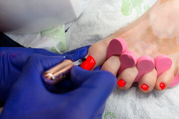 Obraz na płótnie Canvas The manicurist paints hybrid toenails red in blue latex gloves.