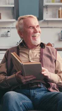 vertical video of smiling elderly man reading book while sitting at home