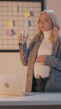vertical video of pregnant businesswoman drinking water and touching belly in office