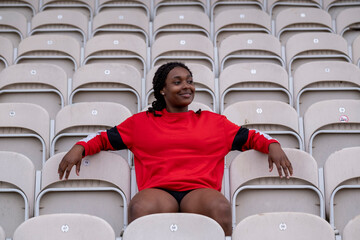 Smiling athletic woman sitting on chair at empty stadium - obrazy, fototapety, plakaty