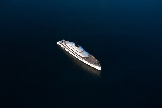 A white large super modern yacht stands on smooth water in the morning rays of the sun aerial view. White large modern yacht on dark water top view.
