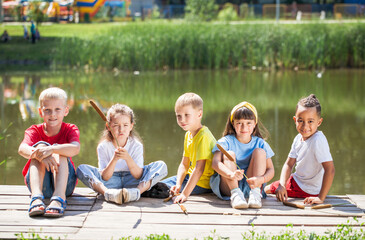 Five happy children sit on a wooden pier near a lake in a summer park
