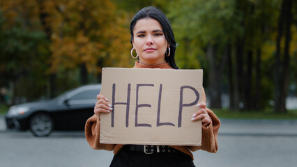 Close-up sad young woman stands outdoors near roadway holding cardboard sign with words HELP upset...