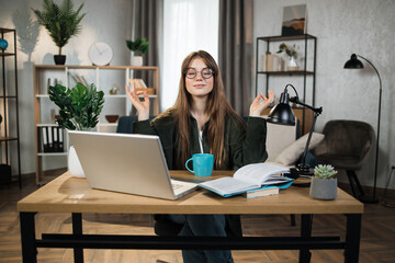 Pretty woman freelancer in eyeglasses sitting at table with closed eyes and relieving stress by...