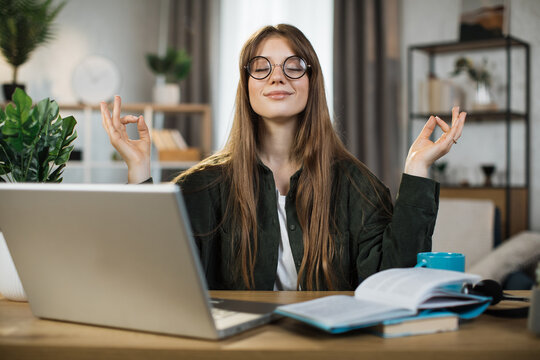 Pretty woman freelancer in eyeglasses sitting at table with closed eyes and relieving stress by meditation at workplace. Concept of relaxation and harmony, no stress free relief at work