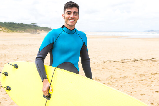 Happy young ethnic guy smiling holding surfboard on seashore