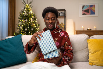 A cheerful man in a warm sweater sits on a couch in a cozy warm living room lit by Christmas tree lights. The man opens the gift box with surprise.