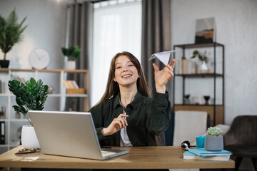 Fototapeta na wymiar Smiling female freelancer playing with paper plane while working from home. Attractive caucasian woman taking break during working time.