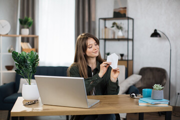 Attractive caucasian woman in casual outfit sitting at desk with modern laptop and playing with paper plane. Female boring at work. Unproductive concept.