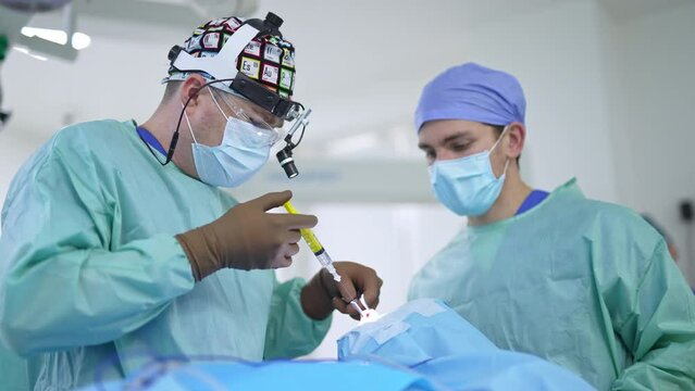 Surgeon in bright cap, mask and device glasses inputs medicines from a syringe into patient’s nose. Male assistant standing near the doctor.