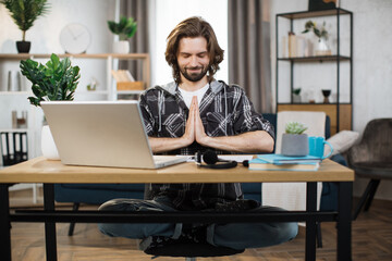 Smiling caucasian man in casual wear sitting on workplace on yoga pose relaxing during break after working with modern laptop. Bearded male freelance meditating with closed eyes.