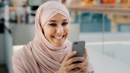 Happy young muslim woman in hijab holding smartphone playing on phone islamic girl smiling relaxes sitting in cafe drinking coffee chatting in social media enjoying using mobile apps for shopping