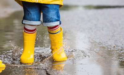 A small child in rainbow socks, yellow rubber boots and a jacket runs through puddles, has fun and...
