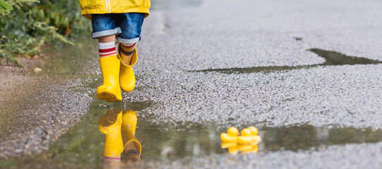 A small child in rainbow socks, yellow rubber boots and a jacket runs through puddles, has fun and plays after the rain. A picture of summer and autumn holidays. Legs close-up. - obrazy, fototapety, plakaty