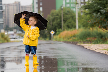 A small child in rainbow socks, yellow rubber boots and a jacket runs through puddles, has fun and plays after the rain. A picture of summer and autumn holidays. A child under an umbrella.