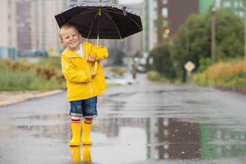 A small child in rainbow socks, yellow rubber boots and a jacket runs through puddles, has fun and plays after the rain. A picture of summer and autumn holidays. A child under an umbrella.