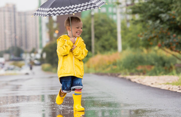 A small child in rainbow socks, yellow rubber boots and a jacket runs through puddles, has fun and plays after the rain. A picture of summer and autumn holidays. A child under an umbrella.