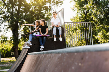 Sports kids with skateboard and penny boards are sitting and chatting with each other on sports ramp on playground. Children communication and friendship. Sports extreme lifestyle.