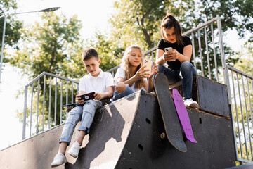 Phone addicted sports kids with skateboard and penny boards are sitting and looking at smartphones on sports ramp on playground. Children addiction of phones.