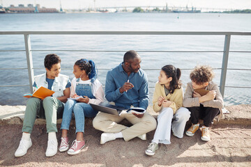 Full length portrait of smiling male teacher with diverse group of children enjoying outdoor class by water in sunlight