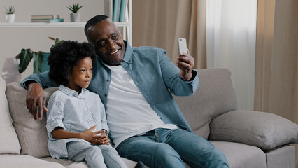 African American family father and daughter sitting in room on couch chatting on webcam mature man with little cute girl shoots video for blog looking at smartphone screen waving hello to phone camera