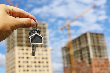 Keychain in house shape and key in male hand on background of construction cranes and new buildings. Real estate agent, concept of buying apartment with a mortgage