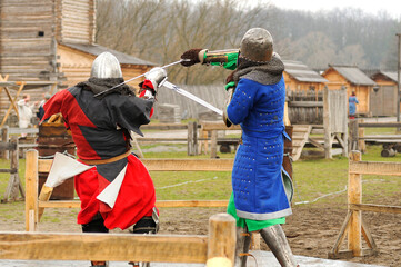 Men reenactors in metal armor of an Old Rus knights reconstructing sword fight, wooden fortress on a background