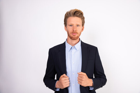 Portrait of serious young businessman looking at camera. Caucasian bearded man wearing blue shirt and black suit coat standing against white background. Successful businessman concept