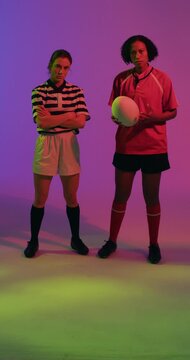 Vertical video of portrait of diverse female rugby players with rugby ball over neon pink lighting