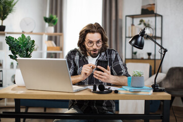 Young bearded excited man in casual outfit typing on mobile phone while sitting at workplace with laptop. Concept of corporate communication and good news.