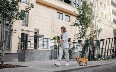 Beautiful Woman wearing white suit and sunglasses walking down the street with Welsh Corgi Pembroke dog - 522283228