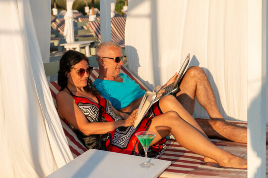 Portrait of a married couple lying on a daybed. Man working on his laptop while his wife is reading a book at the beach. Business and summer vacation - concept.