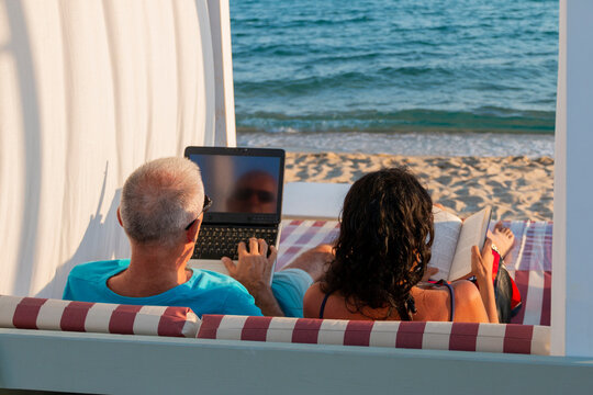 Portrait of a married couple lying on a daybed. Man working on his laptop while his wife is reading a book at the beach. Business and summer vacation - concept.