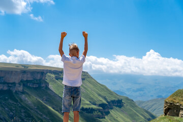 Teenager boy standing on top of mountain with raised hands, looking away with pleasure. School boy hiking in mountains