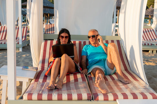 Portrait of a married couple lying on a daybed. Man is on his smartphone, while his wife working on her laptop. Business and summer vacation - concept.