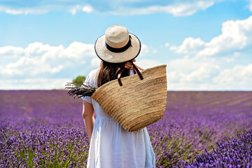 Back view of woman in linen dress and straw hat standing in purple blooming lavender flowers field...