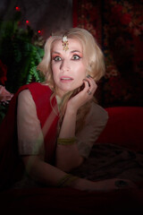 A beautiful European girl looking like an Arab woman in a red room in a harem. Photo shoot of an oriental style odalisque. A model poses in a sari as an indian woman in india